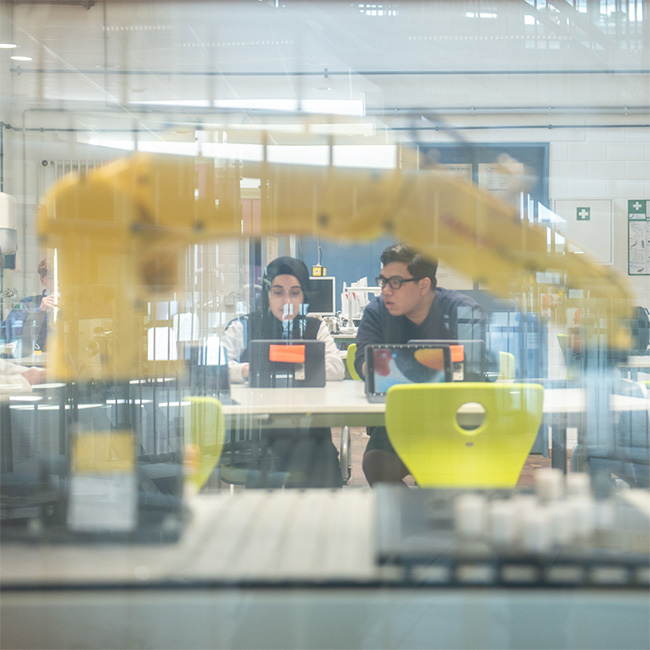 Schülergruppe im Robotiklabor bei der Arbeit mit iPads. Im Vordergrund ein Industrieroboter.